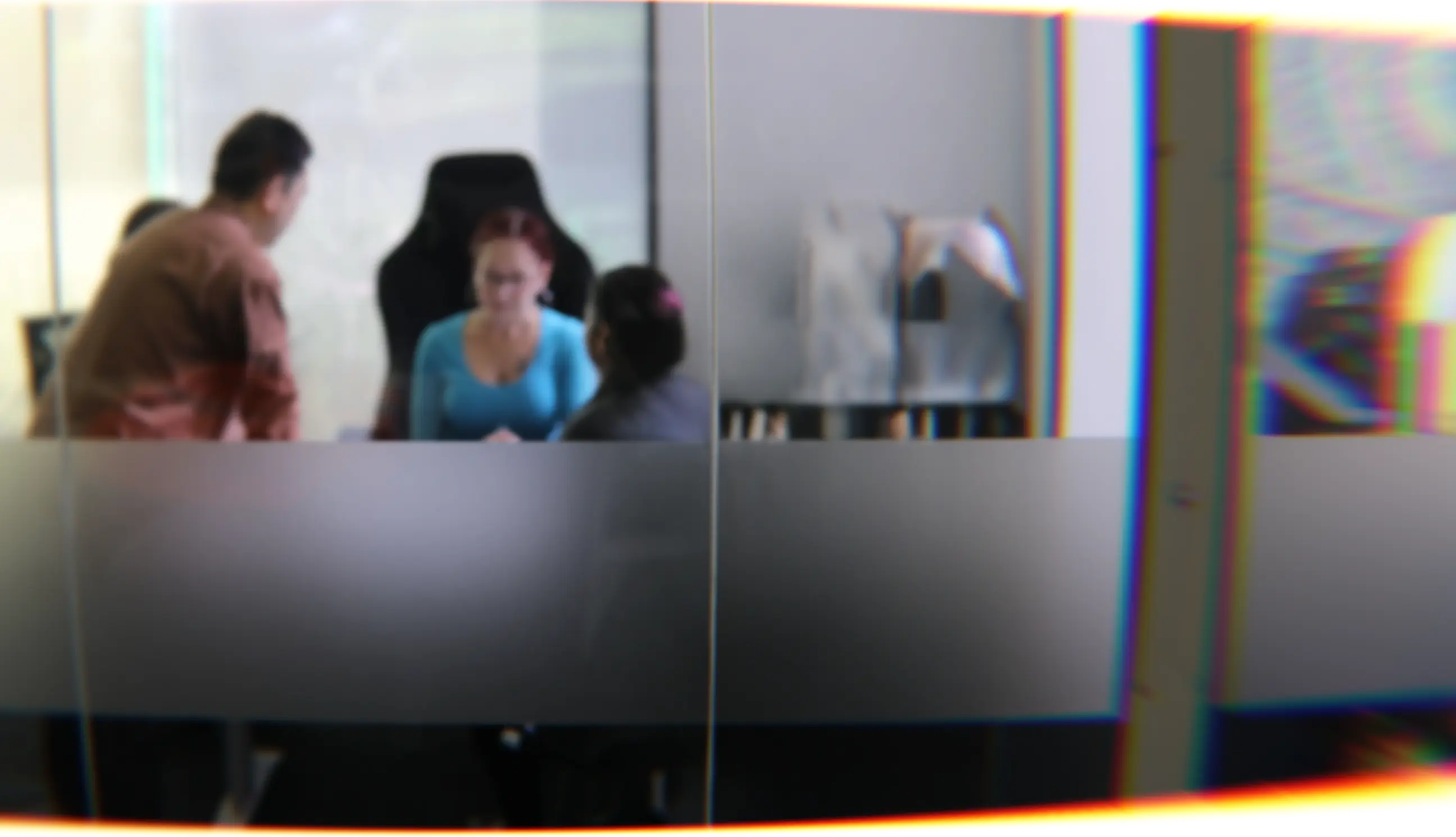 People working on laptops inside a glass-walled office meeting room with a hallway on the left.