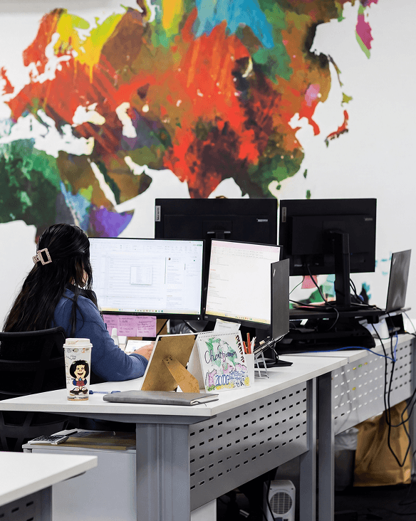 Person working at a desk with dual monitors in an office with a colorful world map mural.